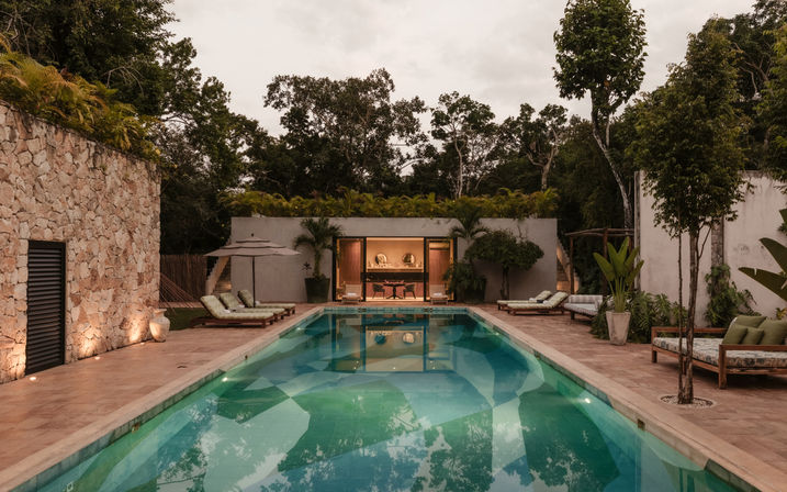 Tropical resort-style villa with a long illuminated outdoor swimming pool, cushioned lounge chairs, umbrellas and a warm-lit open pavilion set among lush trees at dusk.