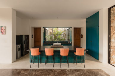 Modern open kitchen with a dark island and cooktop, teal-tiled island base and cabinets, four coral-orange bar stools, stainless fridge, and a window framing a lush stone-walled courtyard