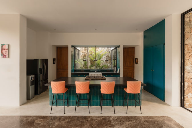 Modern open kitchen with a dark island and cooktop, teal-tiled island base and cabinets, four coral-orange bar stools, stainless fridge, and a window framing a lush stone-walled courtyard