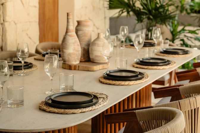 Boho-chic dining table with woven rattan placemats, stacked black plates, wine glasses and water tumblers around a light stone tabletop, accented by rustic ceramic vases and tropical greenery in a stylish restaurant setting.
