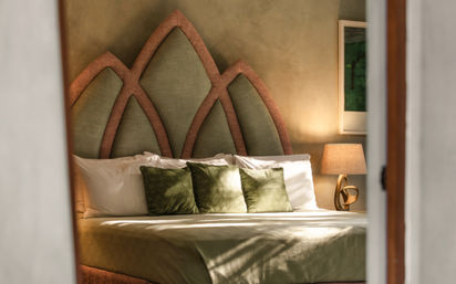 Sun-dappled boutique hotel bedroom with olive-green bedding and three velvet throw pillows, petal-shaped upholstered headboard, bedside lamp and framed art.
