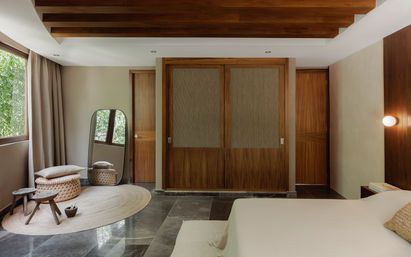 Sunlit modern minimalist bedroom with warm wood beams and sliding wardrobe, marble floor, round jute rug, woven pouf, rustic stools and floor mirror beside a leafy window — tropical retreat vibe.
