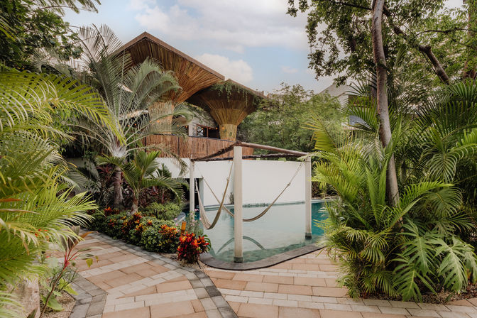 Tropical resort pool scene with a suspended hammock over turquoise water, thatched bamboo canopy, paved walkway and lush palm and tropical foliage