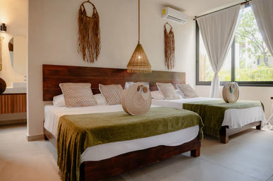 Sunlit modern boho bedroom with two wooden-framed beds, olive-green throws and patterned pillows, woven pendant light and wall hangings, large window with curtains overlooking lush greenery.