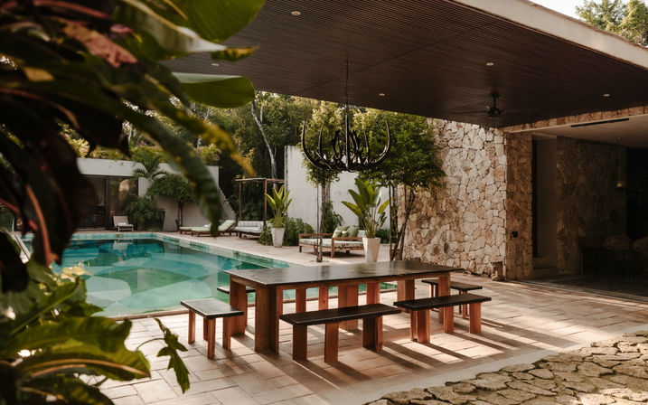 Sunlit tropical poolside patio with a long wooden dining table and benches under a covered terrace, stone wall, hanging chandelier, lounge chairs and lush plants.