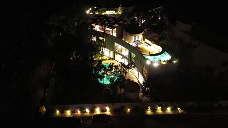 Aerial night view of a modern curved villa with glowing turquoise pools, illuminated terraces and rooftop lounge surrounded by trees and warm pathway lighting.