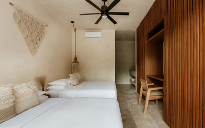 Neutral boho hotel bedroom with two white twin beds, macrame wall hanging, textured woven pillows, hanging wicker lamp, black ceiling fan, wall-mounted AC, tiled floor and wood-slatted closet with desk and chair.