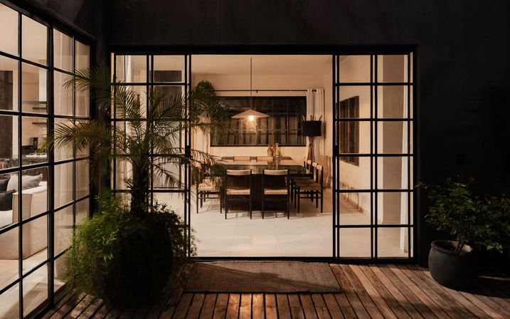 Inviting modern dining room seen through black-framed floor-to-ceiling glass doors from a wooden patio with potted plants and a glowing pendant light.