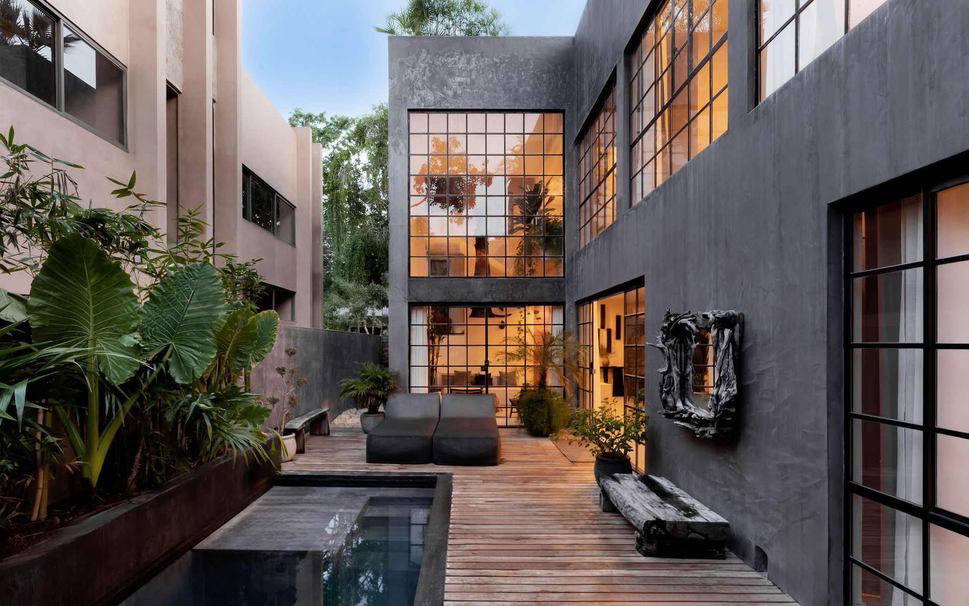 Modern urban courtyard with dark concrete facade and large industrial grid windows glowing warm, wooden deck beside a narrow plunge pool, tropical plants, sculptural wall art and lounge seating.