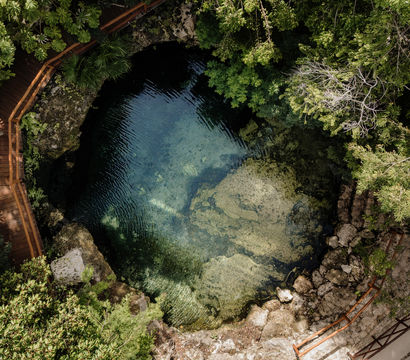 Aerial view of a clear blue cenote — a natural freshwater sinkhole — surrounded by lush tropical forest and winding wooden boardwalks, revealing rocky ledges and underwater rock formations.