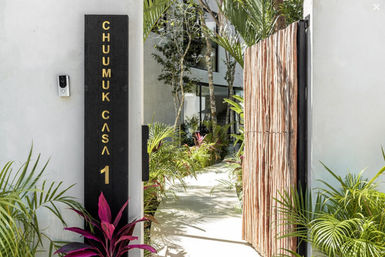 Modern tropical villa entrance with a partially open rustic wooden gate, black vertical address plaque with gold lettering, doorbell camera, and a sunlit concrete path lined with palms and colorful tropical plants leading to a glass-front house.