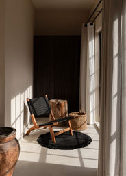 Sunlit cozy reading nook by a tall window with neutral curtains — wooden lounge chair with black woven seat on a round black rug, rustic tree-stump side table and woven basket in a minimalist natural-light interior.
