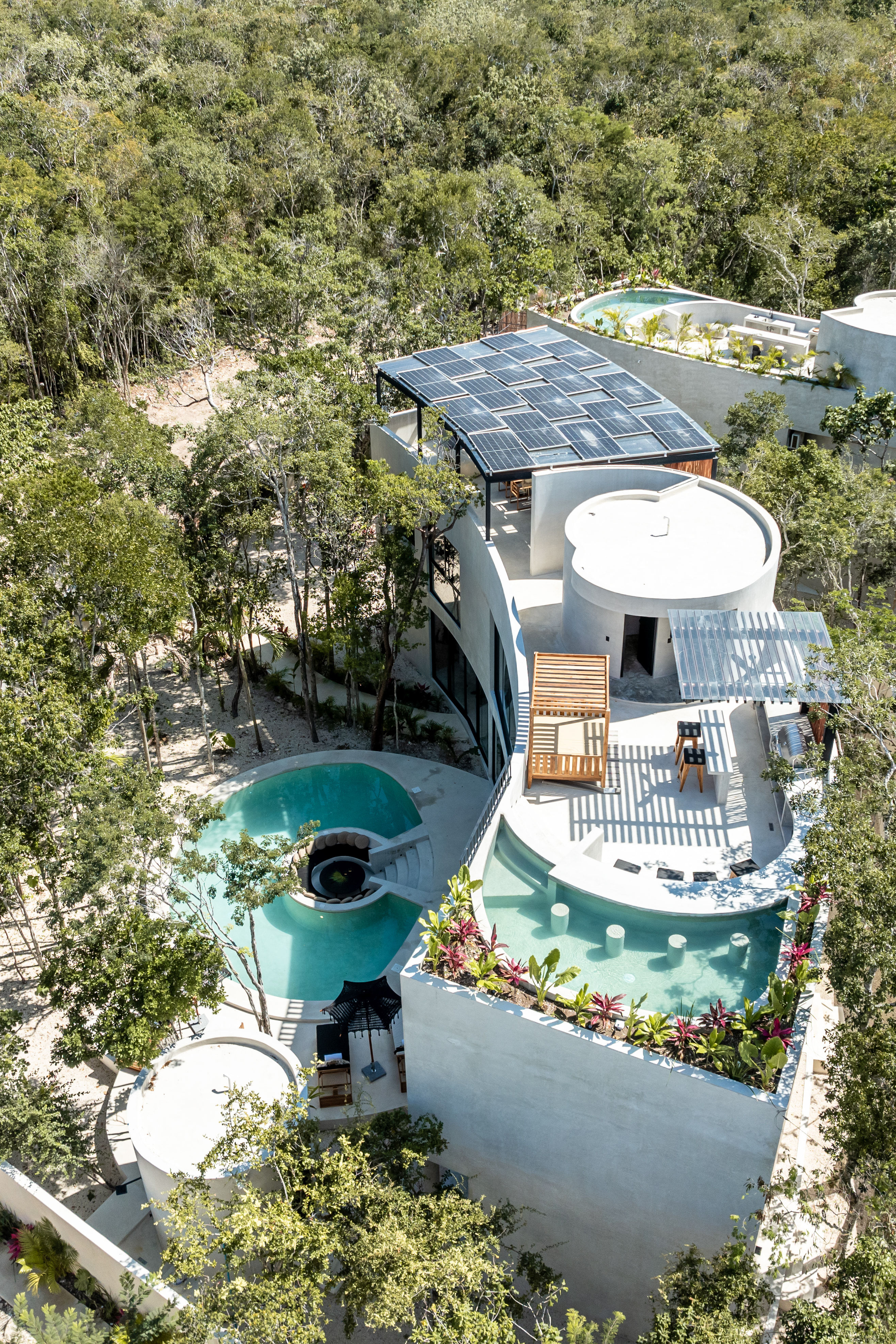 Aerial view of a white curved eco-villa with rooftop solar panels, turquoise winding pools and terraces surrounded by dense tropical forest — sustainable luxury retreat in a jungle setting.
