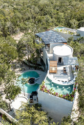 Aerial view of a white curved eco-villa with rooftop solar panels, turquoise winding pools and terraces surrounded by dense tropical forest — sustainable luxury retreat in a jungle setting.