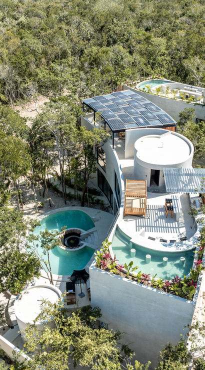 Aerial view of a white curved eco-villa with rooftop solar panels, turquoise winding pools and terraces surrounded by dense tropical forest — sustainable luxury retreat in a jungle setting.