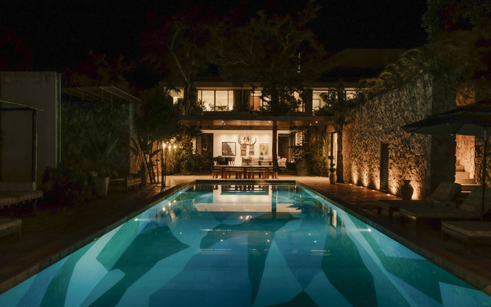 Nighttime view of a luxury tropical villa with a glowing rectangular swimming pool, stone-clad patio, outdoor dining area and lush plants reflected in the water.