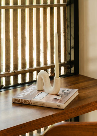 Sunlit wooden table by a slatted window with a modern curved ceramic candleholder holding a tall taper candle, placed on a coffee-table book about wood interiors — minimal warm decor.