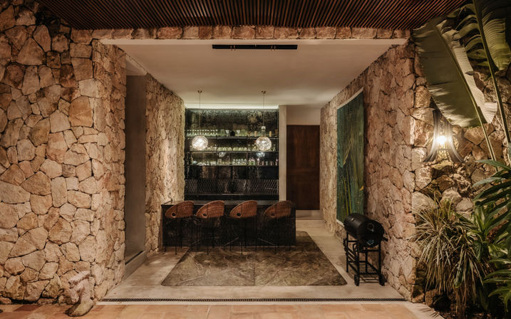 Cozy resort-style outdoor bar set in natural stone walls with four rattan stools, mirrored glass shelving, hanging globe pendant lights, tropical plants, and a small grill under warm evening lighting.