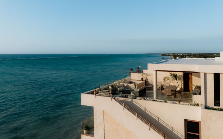 Sunlit oceanfront rooftop terrace with glass‑railed balcony, outdoor dining table and lounge seating overlooking turquoise sea and clear blue sky