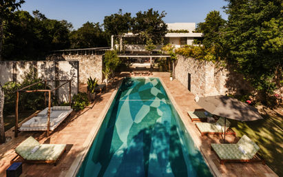 Sunlit long lap pool at a luxury tropical villa with turquoise mosaic water, terracotta decks, stone retaining walls, cushioned daybeds, umbrella and a modern white house framed by lush trees.