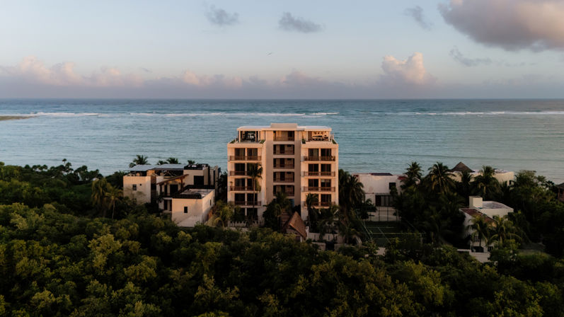 Beachfront mid-rise condominium framed by palm trees and lush tropical greenery, overlooking calm turquoise ocean and a sun‑kissed pastel sky at sunset.