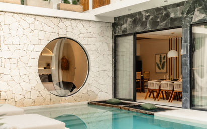 Sunlit modern villa courtyard with turquoise private pool, circular porthole window set in textured white stone wall and sliding glass doors opening to a stylish indoor dining area.