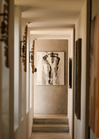 Sunlit minimalist hallway interior with concrete steps leading to a wall-mounted black-and-white abstract painting, neutral plaster walls and decorative brass sconces