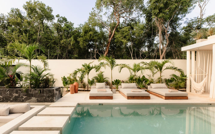Sun-soaked modern poolside patio with three cushioned wooden daybeds, tropical palm plants, terracotta pots and a hammock against a white privacy wall