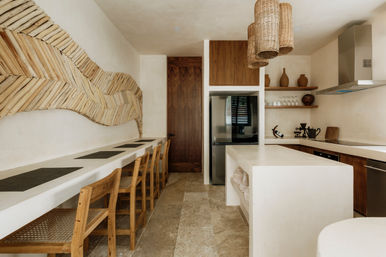 Neutral-toned Mediterranean-inspired kitchen design with a long white breakfast bar and wooden chairs, woven driftwood wall art, wicker pendant lights, central island, stainless refrigerator and open shelves with clay vases