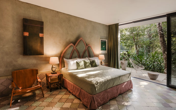 Cozy sunlit boutique-style bedroom with decorative arched headboard, neutral linens, bedside lamps and wooden lounge chair on patterned tile; sliding glass doors open to a lush tropical forest terrace with outdoor seating.