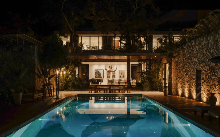 Nighttime view of a modern two-story villa with glass façade and stone walls, warm interior lighting and chandelier, outdoor dining and lush tropical landscaping, and a glowing rectangular swimming pool reflecting the lights.