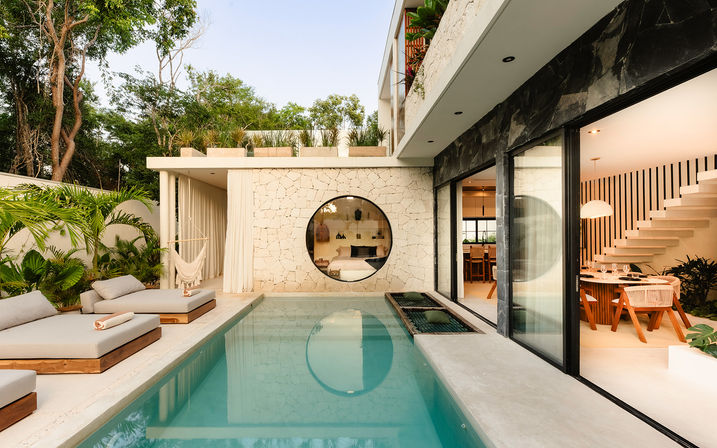 Modern tropical villa patio with a private rectangular pool, cushioned wooden sun loungers, hammock, circular porthole window in a stone wall, and sliding glass doors opening to a dining area and staircase surrounded by lush greenery.