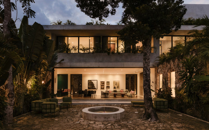 Two-story modern tropical villa at dusk with illuminated open-plan living room, stone courtyard and circular fountain, outdoor lounge chairs, and lush palm landscaping.