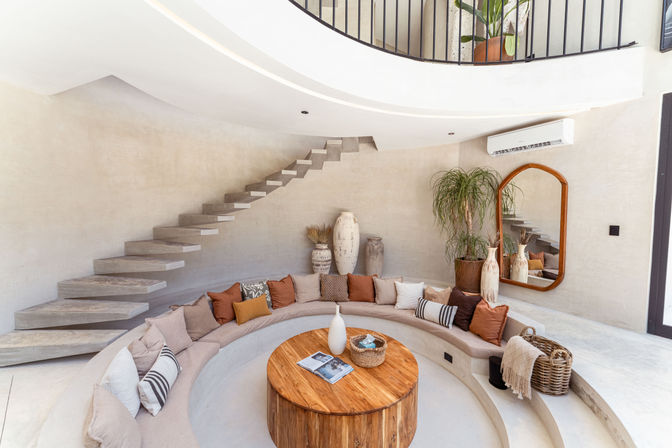Modern sunken living room with curved built-in sofa and rust-beige pillows, round wooden coffee table, floating concrete staircase, large wooden-framed mirror, potted plants and terracotta vases under a white curved mezzanine — airy boho-minimal interior.