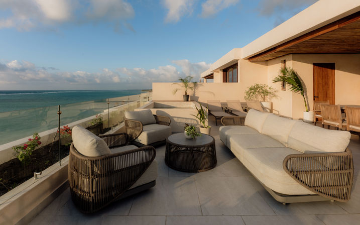Sunlit oceanfront rooftop terrace with modern woven lounge sofa and armchairs around a round coffee table, glass railing, potted palms and sun loungers overlooking turquoise sea and blue sky