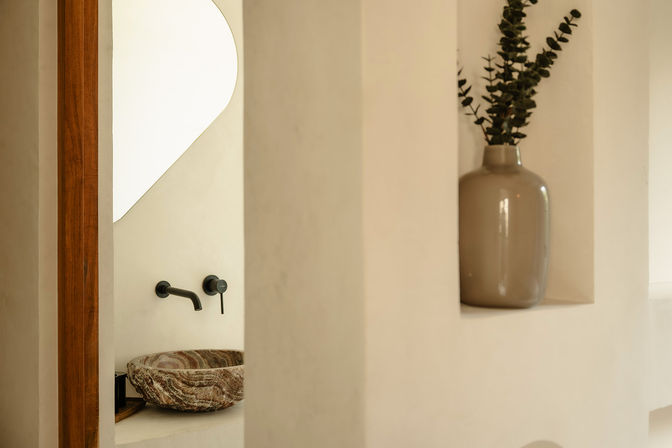 Minimalist spa-like bathroom featuring a carved stone vessel sink, matte black wall faucet, smooth cream plaster walls, warm wood trim, and a recessed niche with a neutral ceramic vase and eucalyptus.