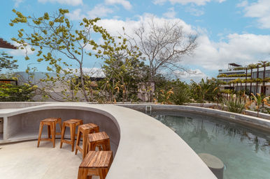 Sunny tropical rooftop poolside with curved concrete bar, wooden stools, lush trees and modern balcony planters under a blue sky with fluffy clouds