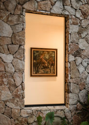 Rustic stone wall window revealing a framed painting of three dogs on a warm cream interior wall, with leafy plants peeking into the foreground.