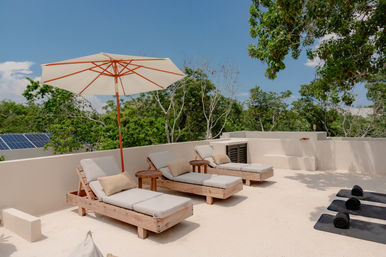 Sun-drenched rooftop terrace with three wooden sun loungers and neutral cushions under a large white parasol, rolled black yoga mats on the deck, solar panels on a low wall and lush green treetops under a clear blue sky.