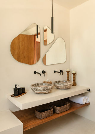 Neutral modern bathroom with a floating concrete double vanity, two stone vessel sinks, matte black wall-mounted faucets, three irregular geometric mirrors, a slim black pendant light, wooden shelf with wicker baskets and rolled towel — spa-like organic design.