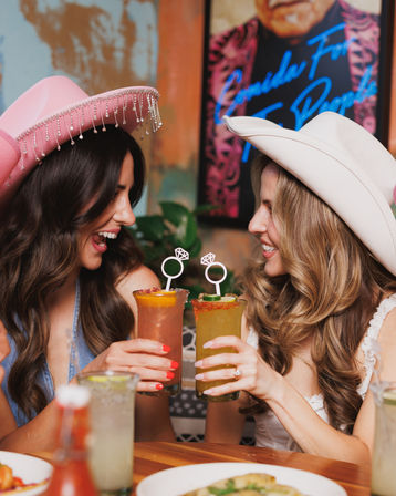 Two friends in pink and white cowgirl hats laughing and clinking colorful cocktails with ring-shaped stirrers at a lively brunch