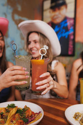 Two friends in cowboy hats clinking colorful cocktails topped with diamond-ring stirrers over tacos on a wooden table — festive restaurant celebration