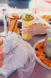 Hand holding a jalapeño-topped mango margarita in a copper-rim glass with lime slices and rose-gold straw, beside a fruit cocktail and pizza on a brunch table.