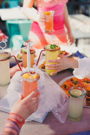 Sunny outdoor patio brunch scene of friends toasting vibrant citrus cocktails garnished with lime and jalapeño, tall glasses with playful ring-shaped stirrers and shared pizza on the table.
