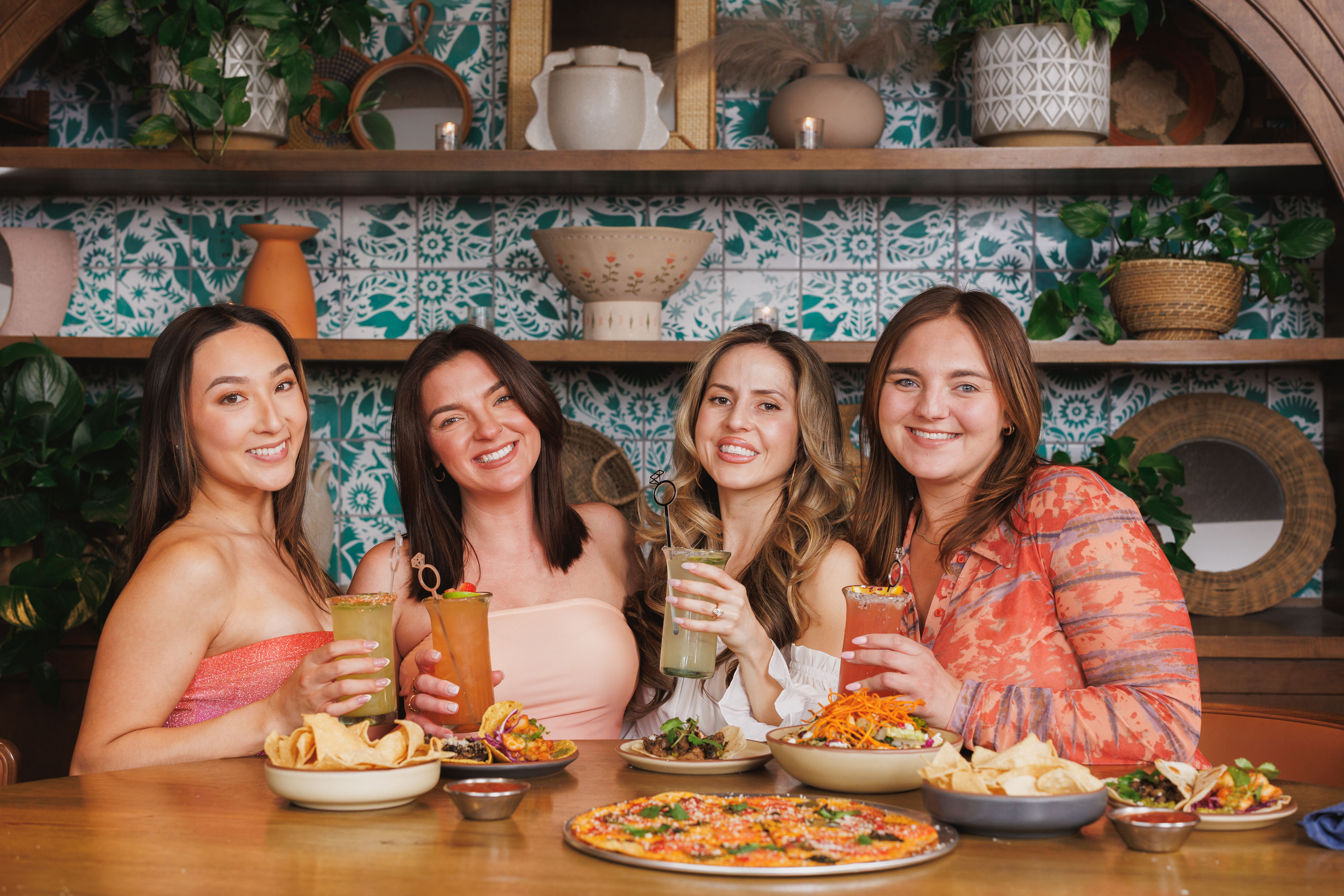 Four friends toasting colorful cocktails over pizza, tacos and chips at a cozy indoor restaurant with teal patterned tile and decorative shelves — cheerful group dining scene.