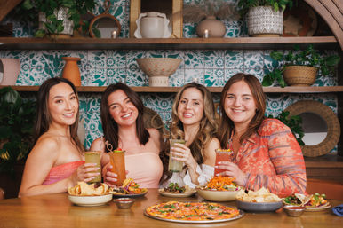 Four friends toasting colorful cocktails over pizza, tacos and chips at a cozy indoor restaurant with teal patterned tile and decorative shelves — cheerful group dining scene.