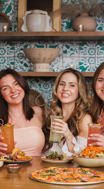 Four friends toasting colorful cocktails over pizza, tacos and chips at a cozy indoor restaurant with teal patterned tile and decorative shelves — cheerful group dining scene.