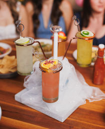 Tall fruity cocktails on a wooden table with salted rims, citrus garnishes and ring-shaped stirrers, blurred friends in the background — festive brunch drinks.