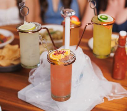 Tall fruity cocktails on a wooden table with salted rims, citrus garnishes and ring-shaped stirrers, blurred friends in the background — festive brunch drinks.