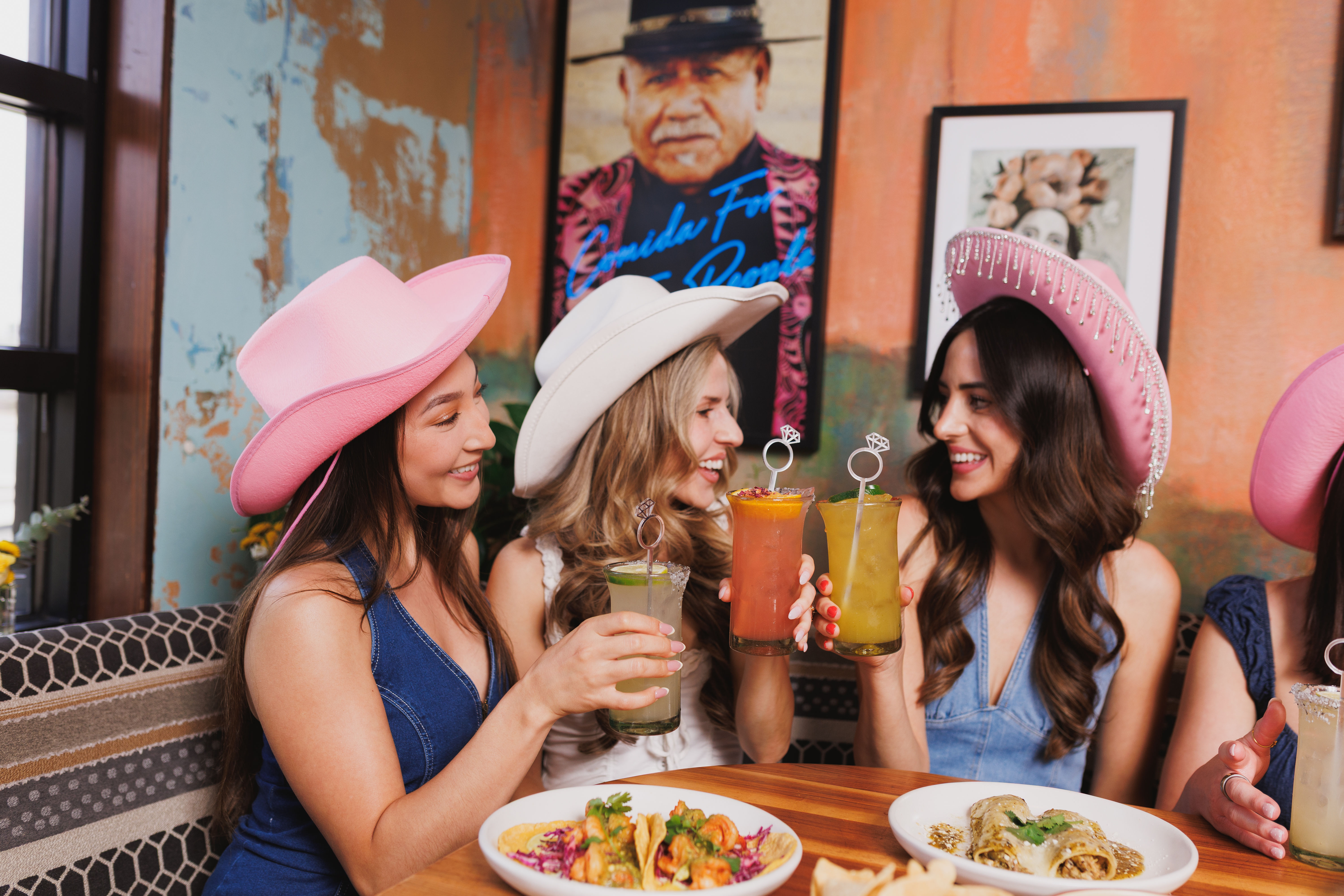 Three women in pink and white cowboy hats cheerfully clinking colorful cocktails over tacos and enchiladas at a lively Mexican-style restaurant